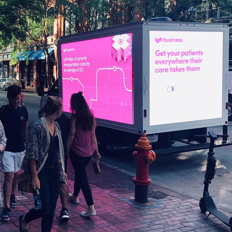 People walking beside a mobile LED billboard displaying vibrant advertisements.